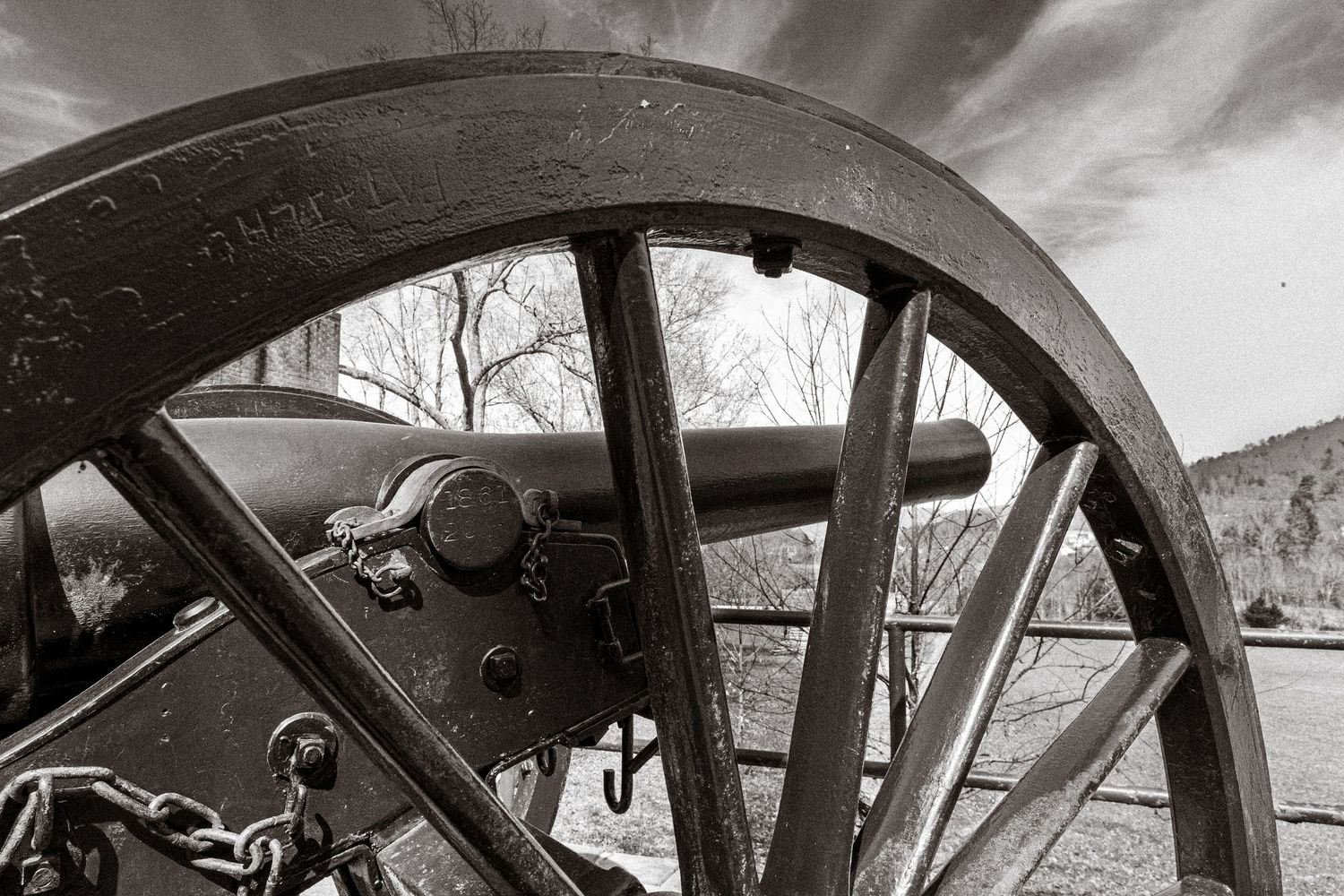 Black and white photo of a Civil War cannon framed by a large wooden wagon wheel spoke at a historic battlefield.