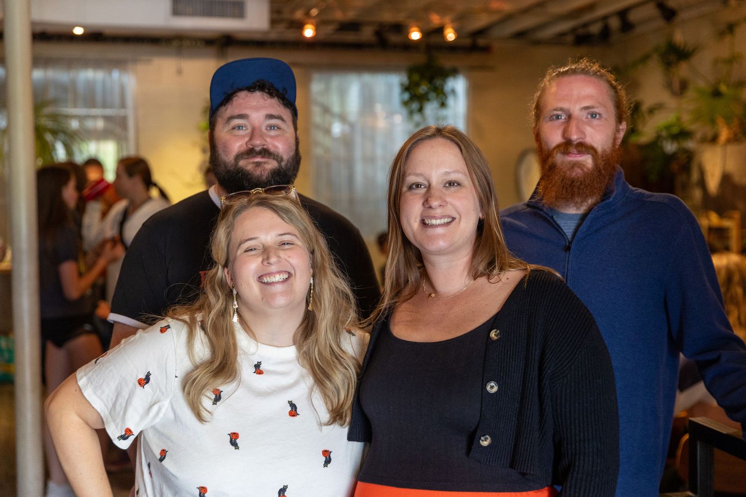 Four smiling friends pose together indoors at a casual social gathering, two women in front and two bearded men behind.