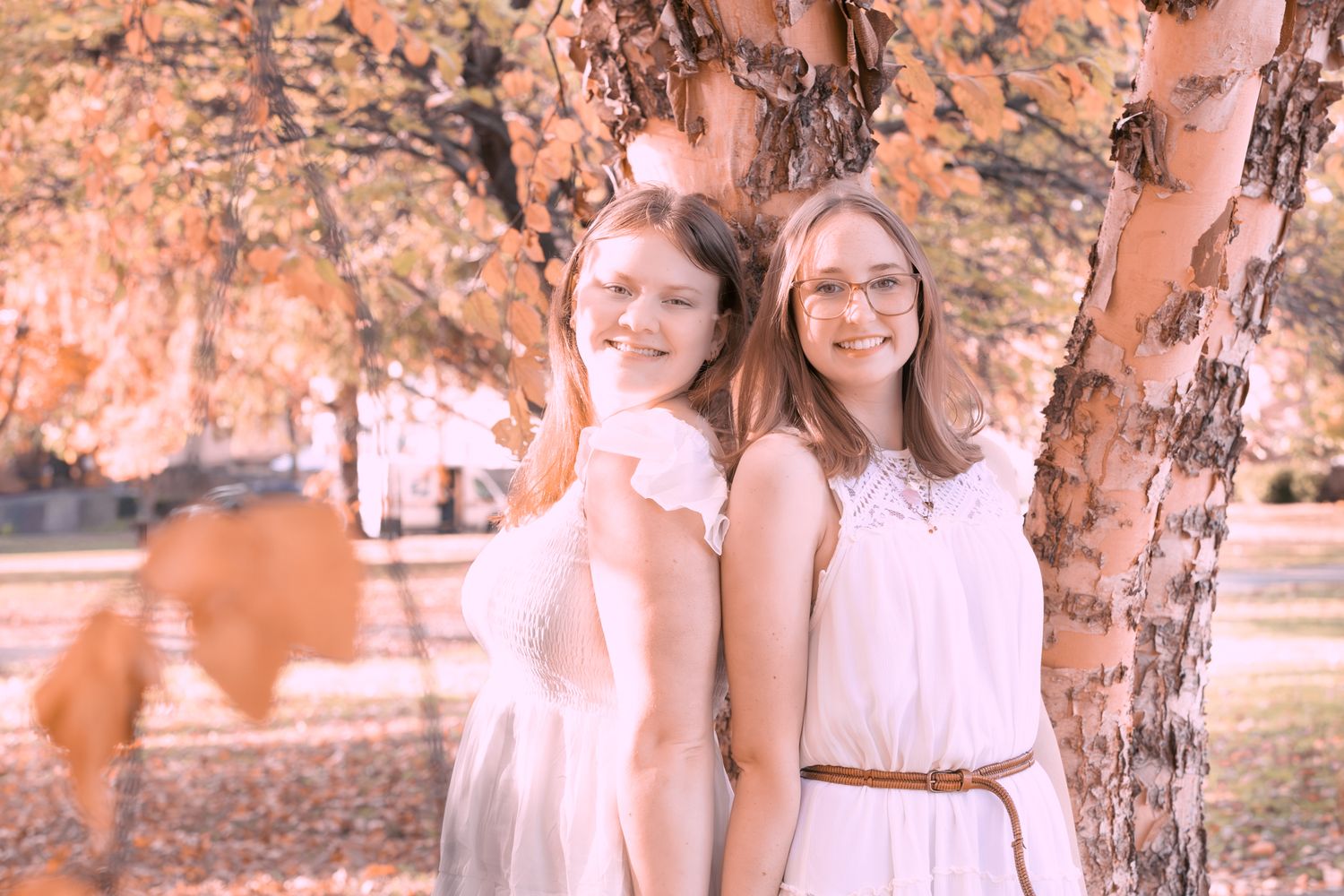 A group of people in white outfits pose together under autumn trees with orange and peach tones in the background.