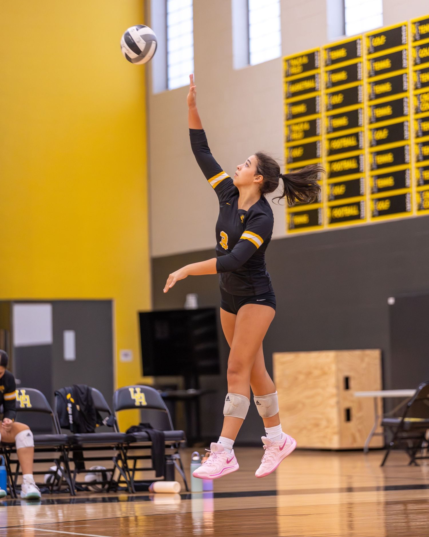 Volleyball player in black and gold uniform jumps to serve the ball during an indoor gym match.
