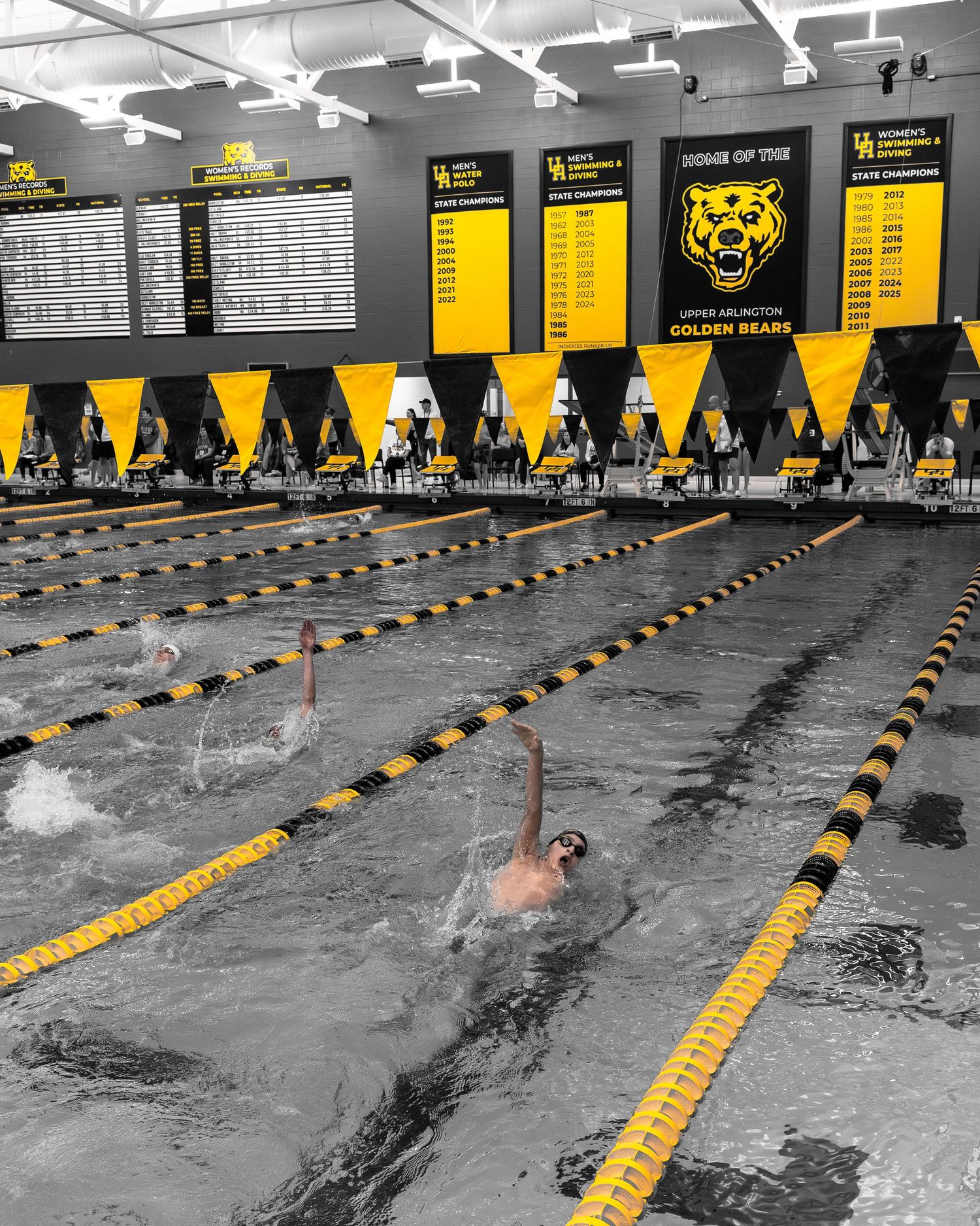 Swimmers race in an indoor pool at Upper Arlington Golden Bears natatorium with yellow lane lines and championship banners.