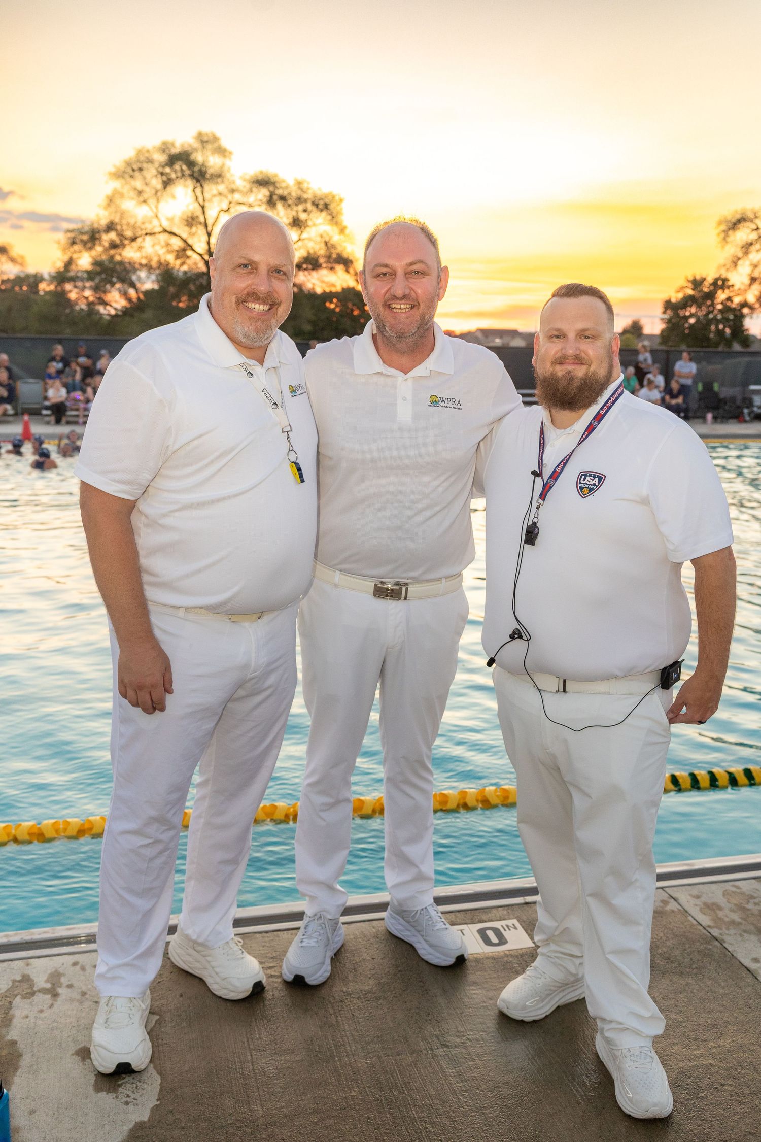 Three men in white uniforms pose poolside at sunset during an outdoor swimming event.
