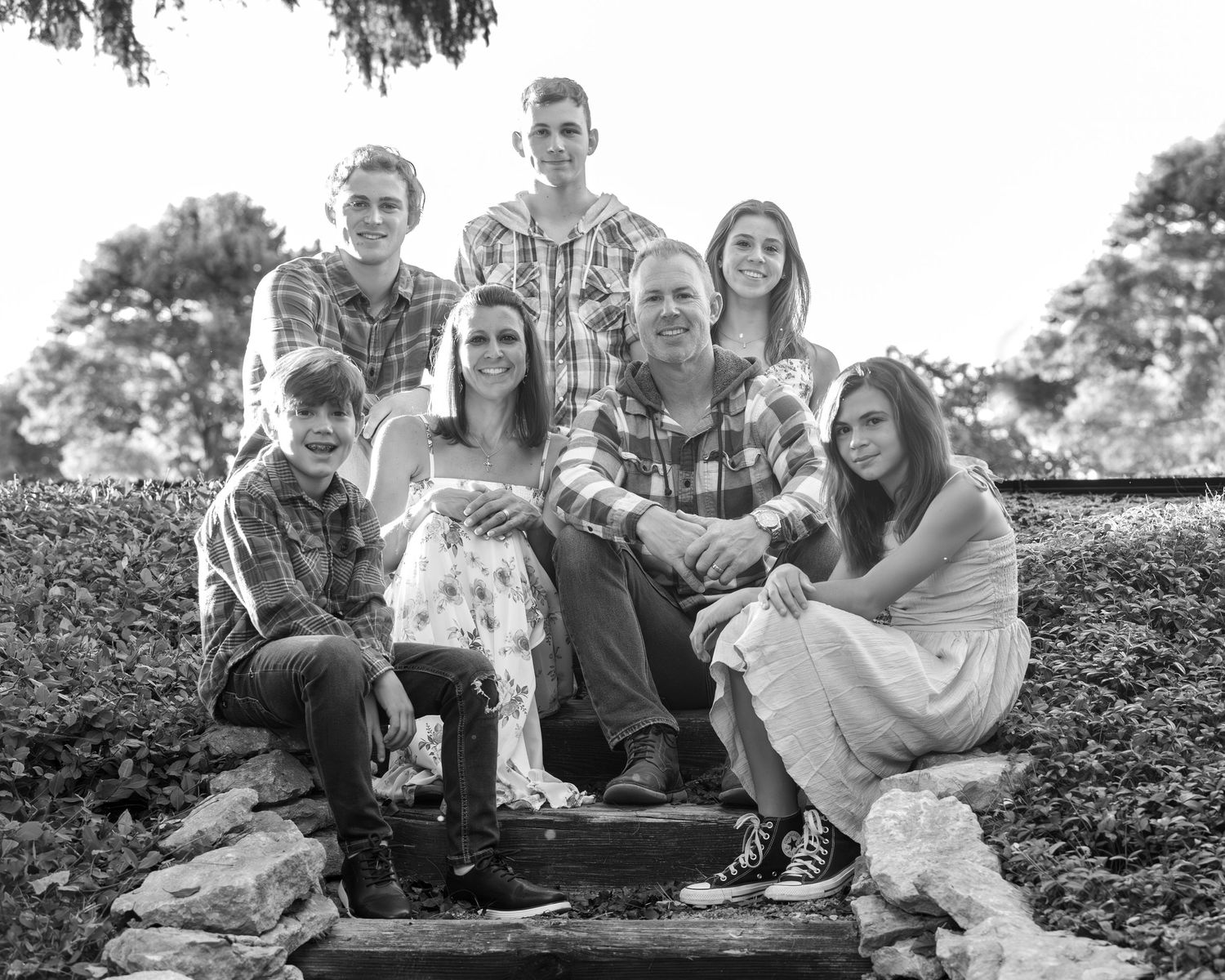Black and white family portrait of seven people posing outdoors on stone steps surrounded by lush greenery.