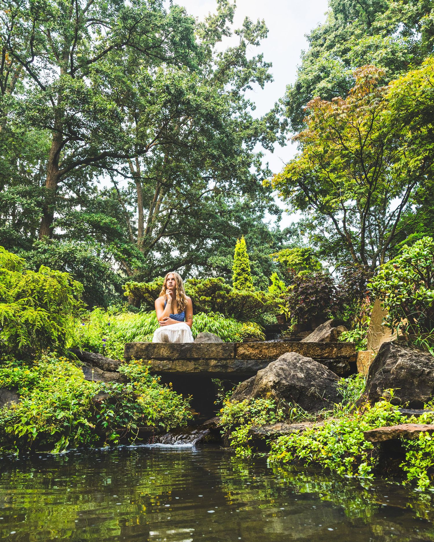 Woman sitting on rocks beside a tranquil pond surrounded by lush green trees in a serene Japanese garden.