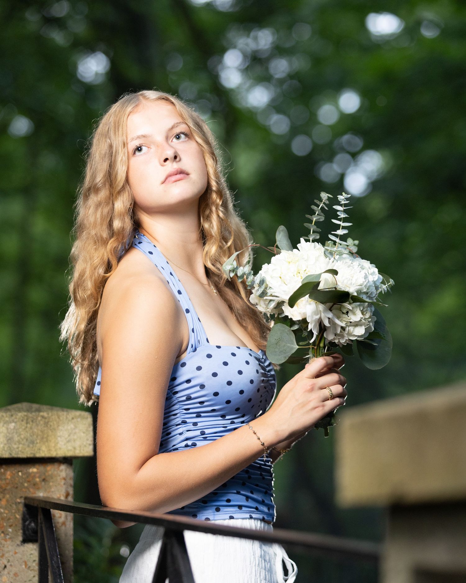 Young blonde woman in blue polka dot dress holding white flower bouquet, gazing upward outdoors.