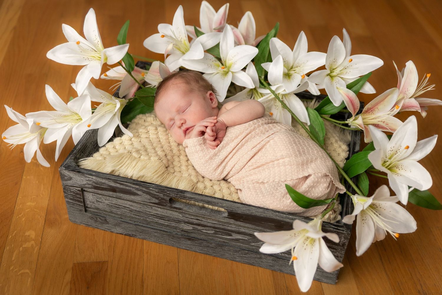 Newborn baby sleeping peacefully in a rustic wooden crate surrounded by white lily flowers on a wooden floor.