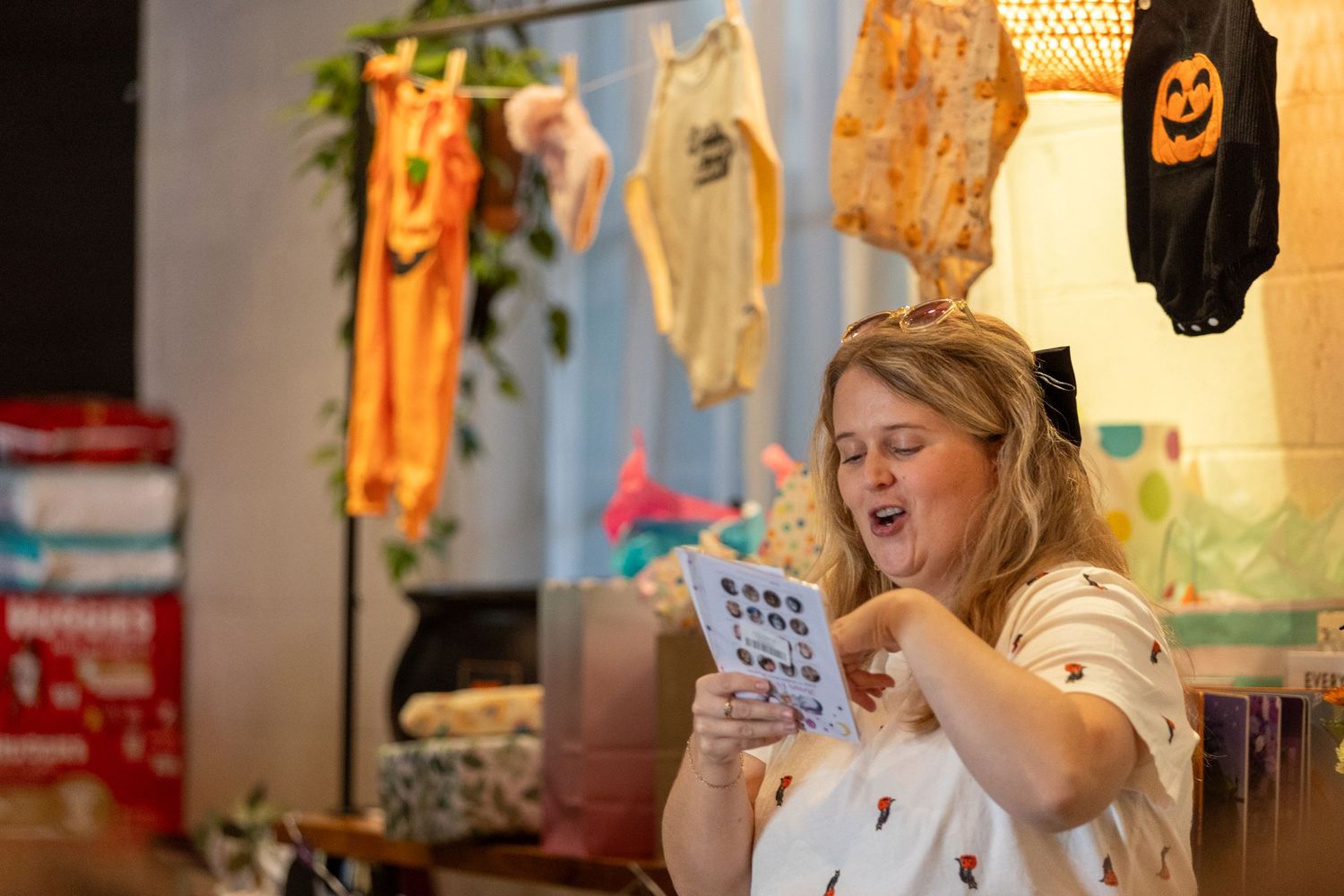 Woman laughing at baby shower bingo card with colorful baby clothes hanging on display in festive decorated room.