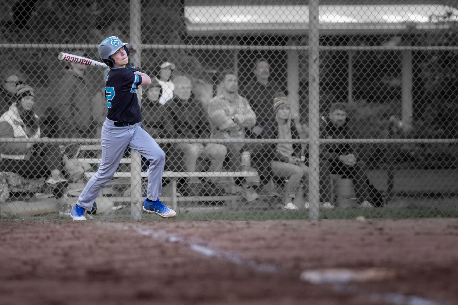 Young baseball player swinging bat at home plate during a little league game, with spectators watching behind chain-link fence.