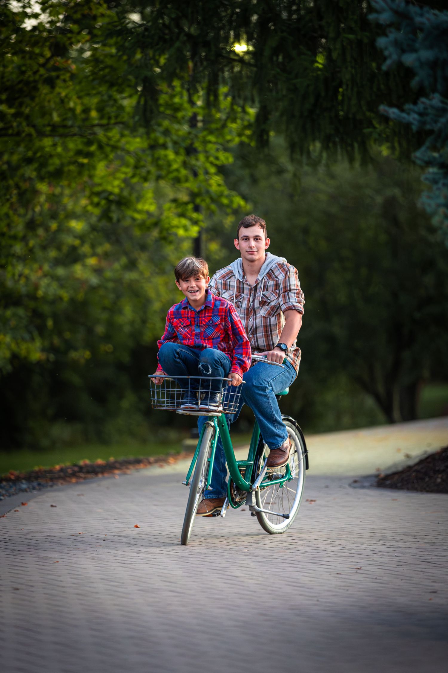A father and son ride a green bicycle together on a park path, both wearing plaid shirts, smiling joyfully.