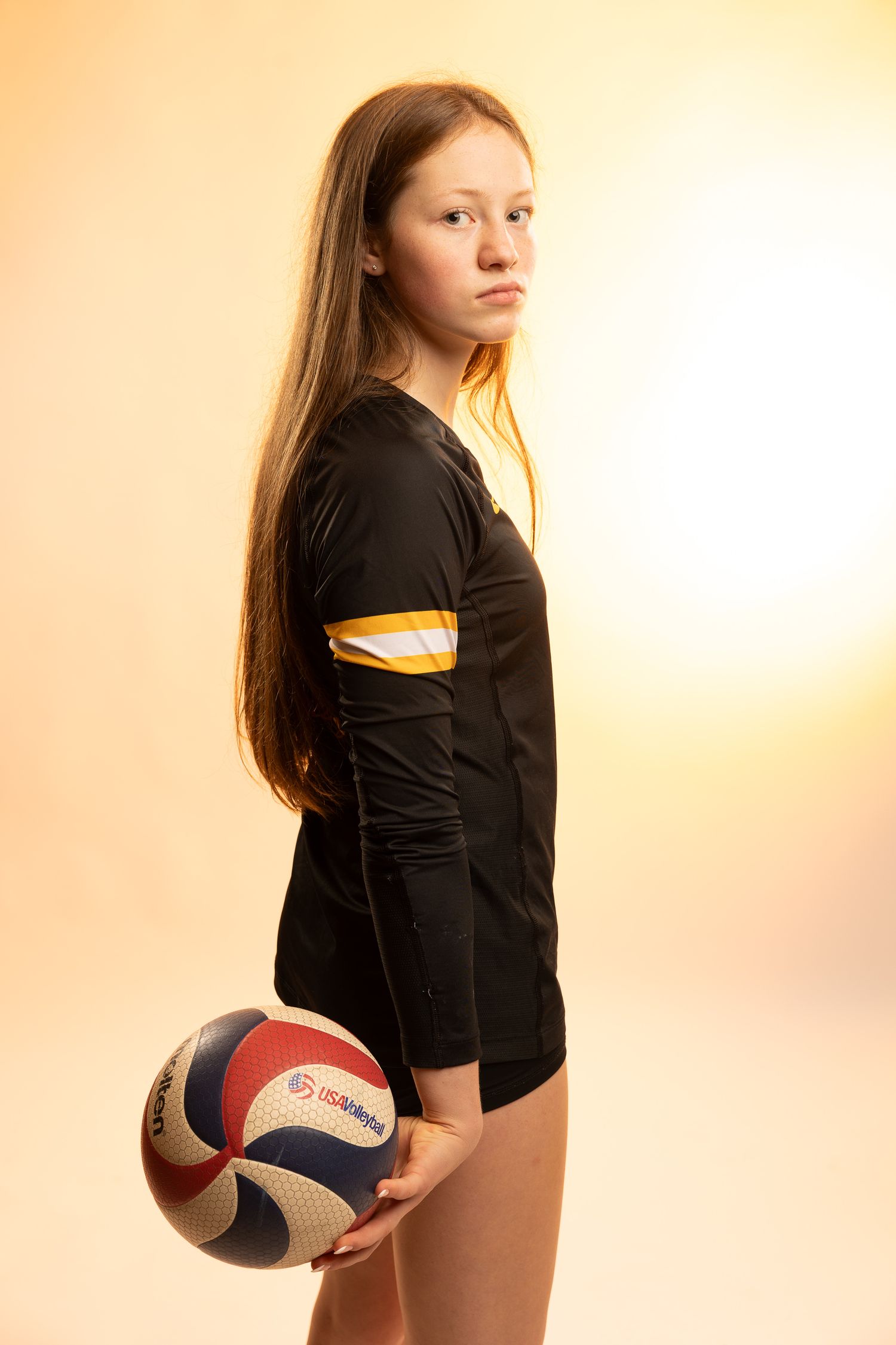 Young female volleyball player in black jersey holds a volleyball, posing seriously against a warm golden background.
