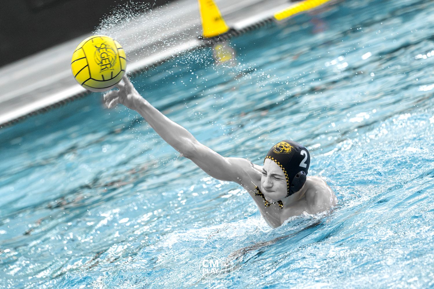 Water polo player in yellow cap reaching to catch a ball during a competitive swim match in an indoor pool.