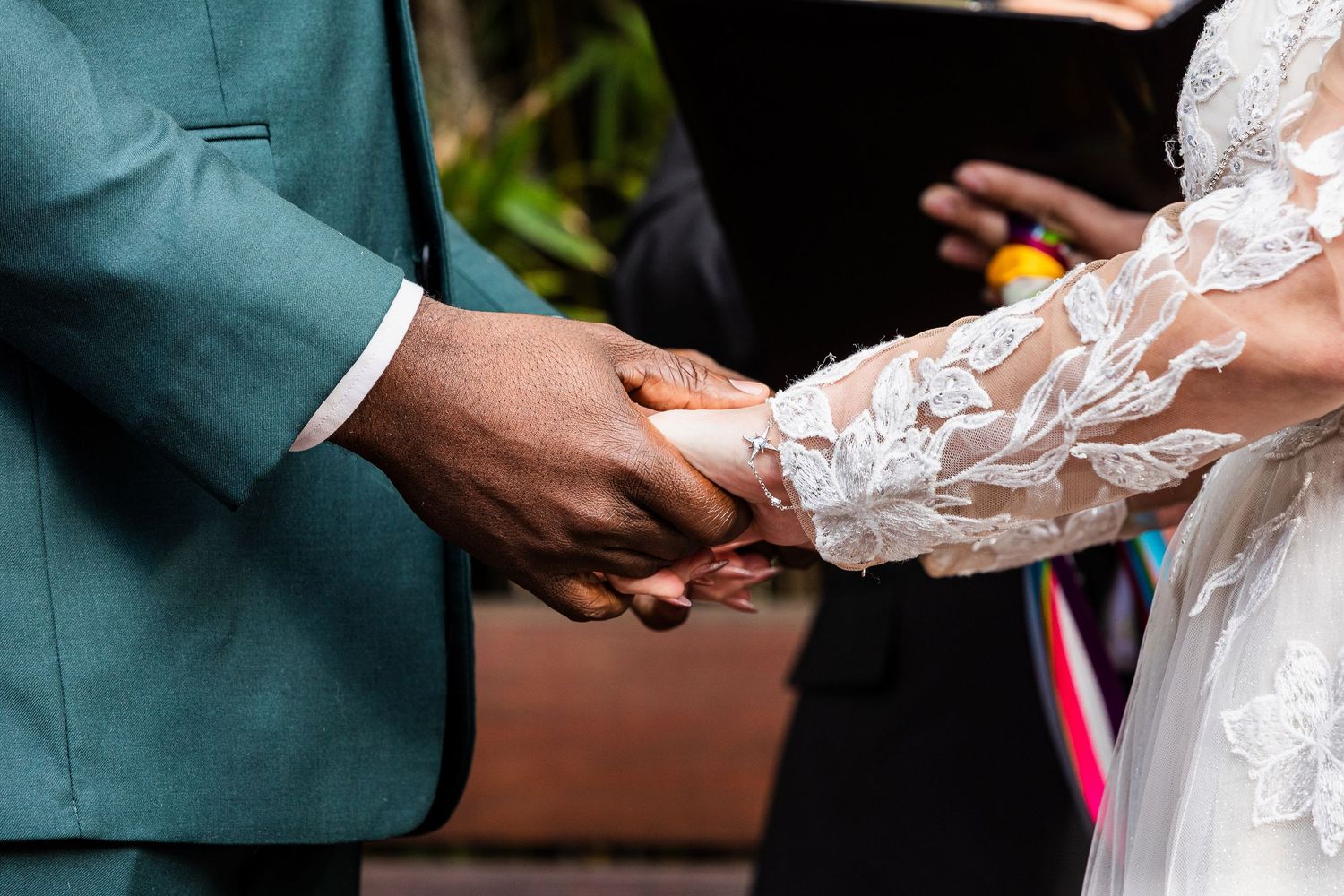 Close-up of hands holding during wedding ceremony with green suit and white lace dress details.