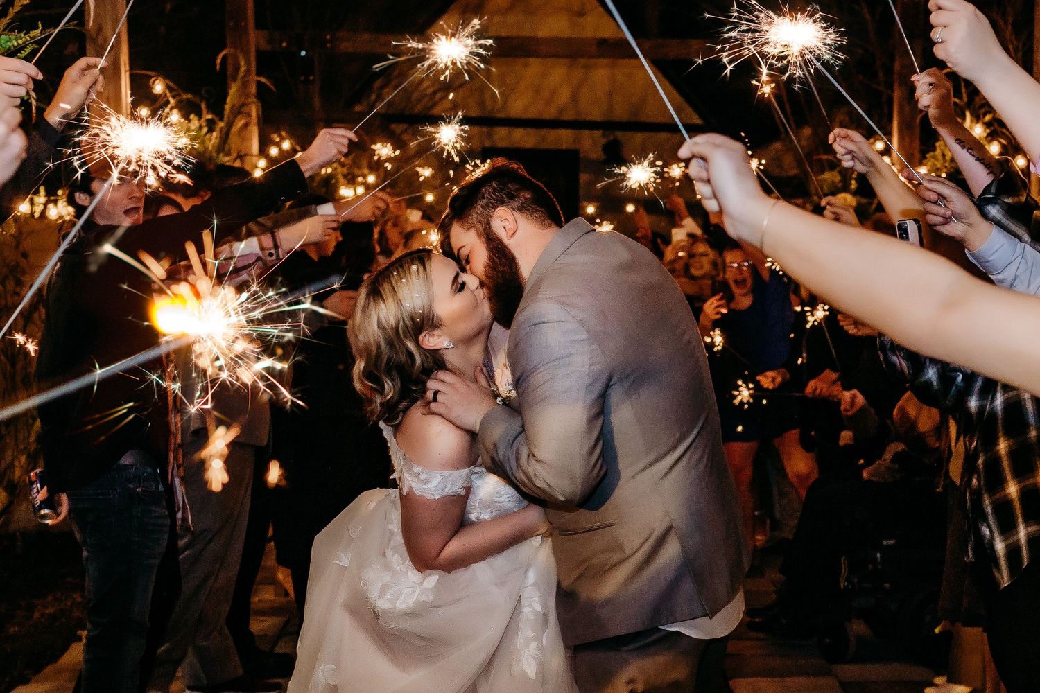 A newlywed couple shares a romantic kiss under sparklers held by wedding guests during their evening celebration.