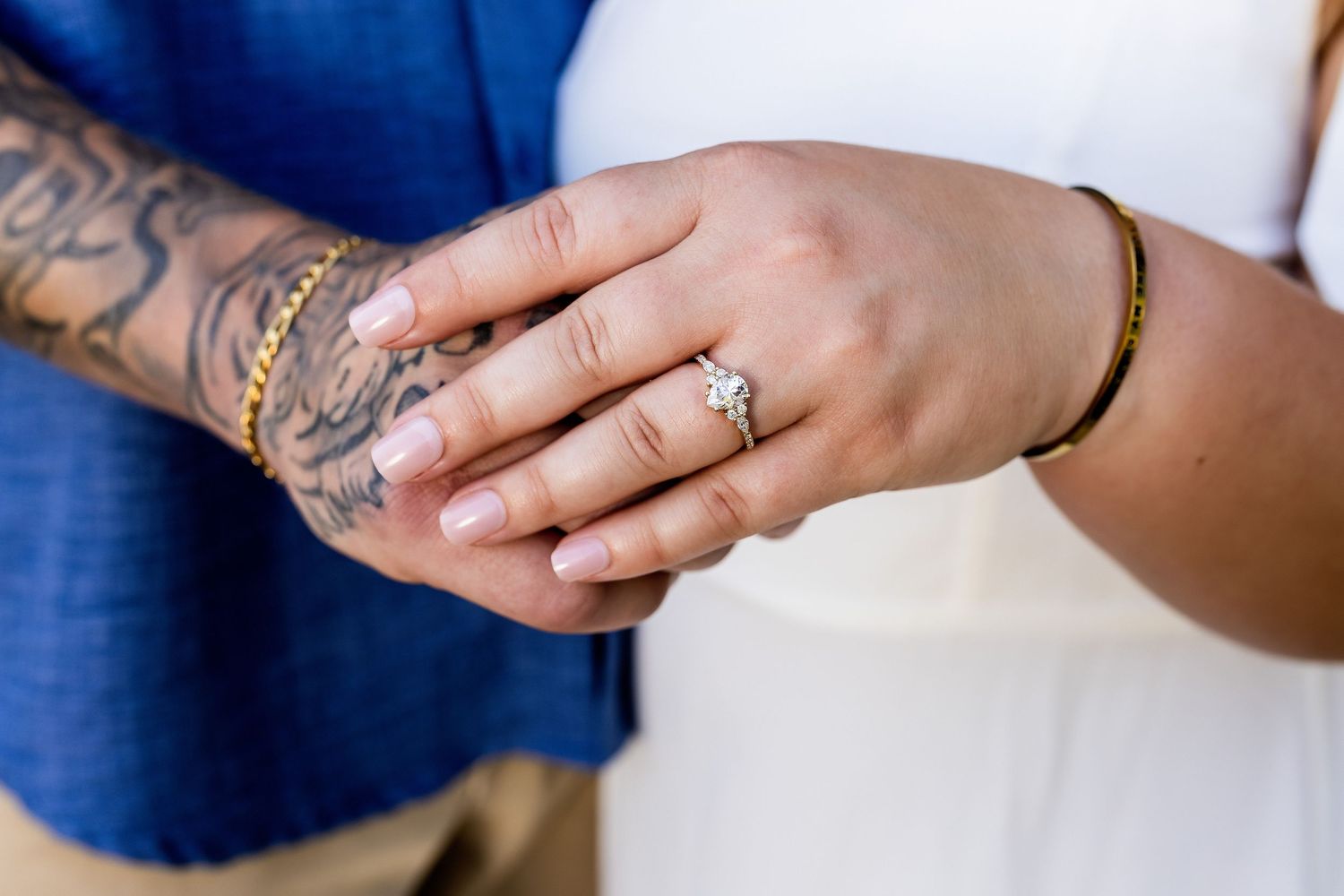 Close-up of hands displaying an engagement ring and intricate arm tattoo against white and blue clothing.