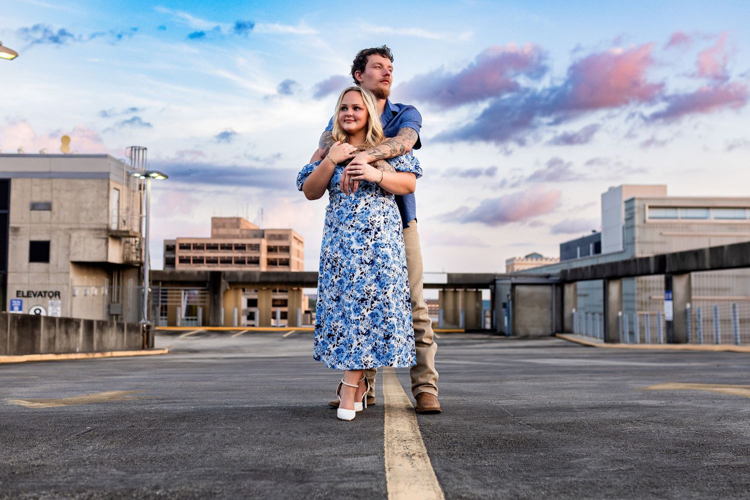 A couple embracing on a rooftop during sunset with cityscape and colorful sky in the background.