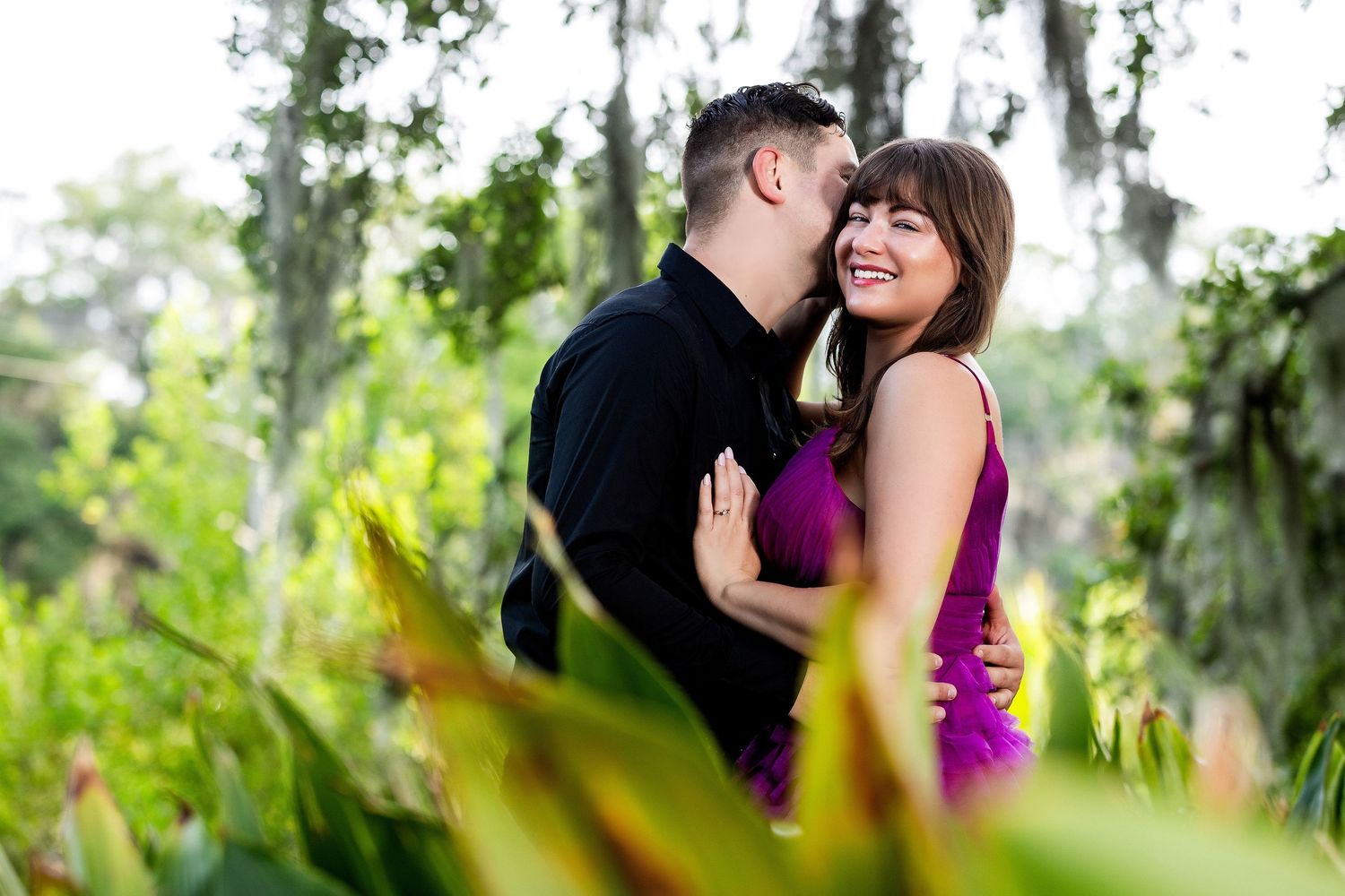 A romantic couple embraces and laughs together in a lush garden setting with Spanish moss and greenery.