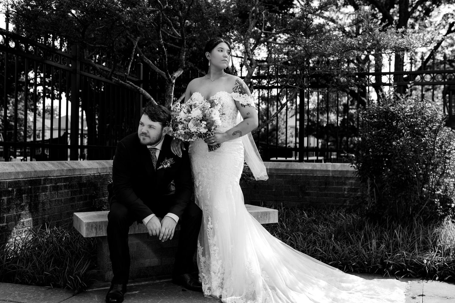 A black and white wedding photo showing a couple in formal attire posing in front of a wrought iron gate outdoors.
