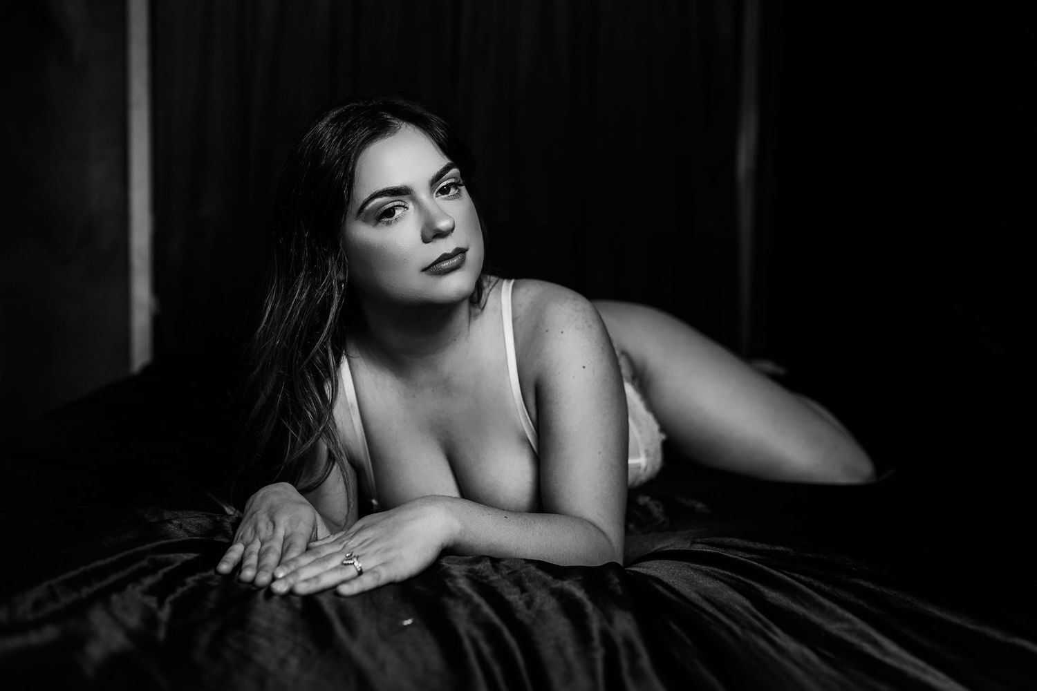 A dramatic black and white boudoir portrait of a person lying on a bed in moody lighting.