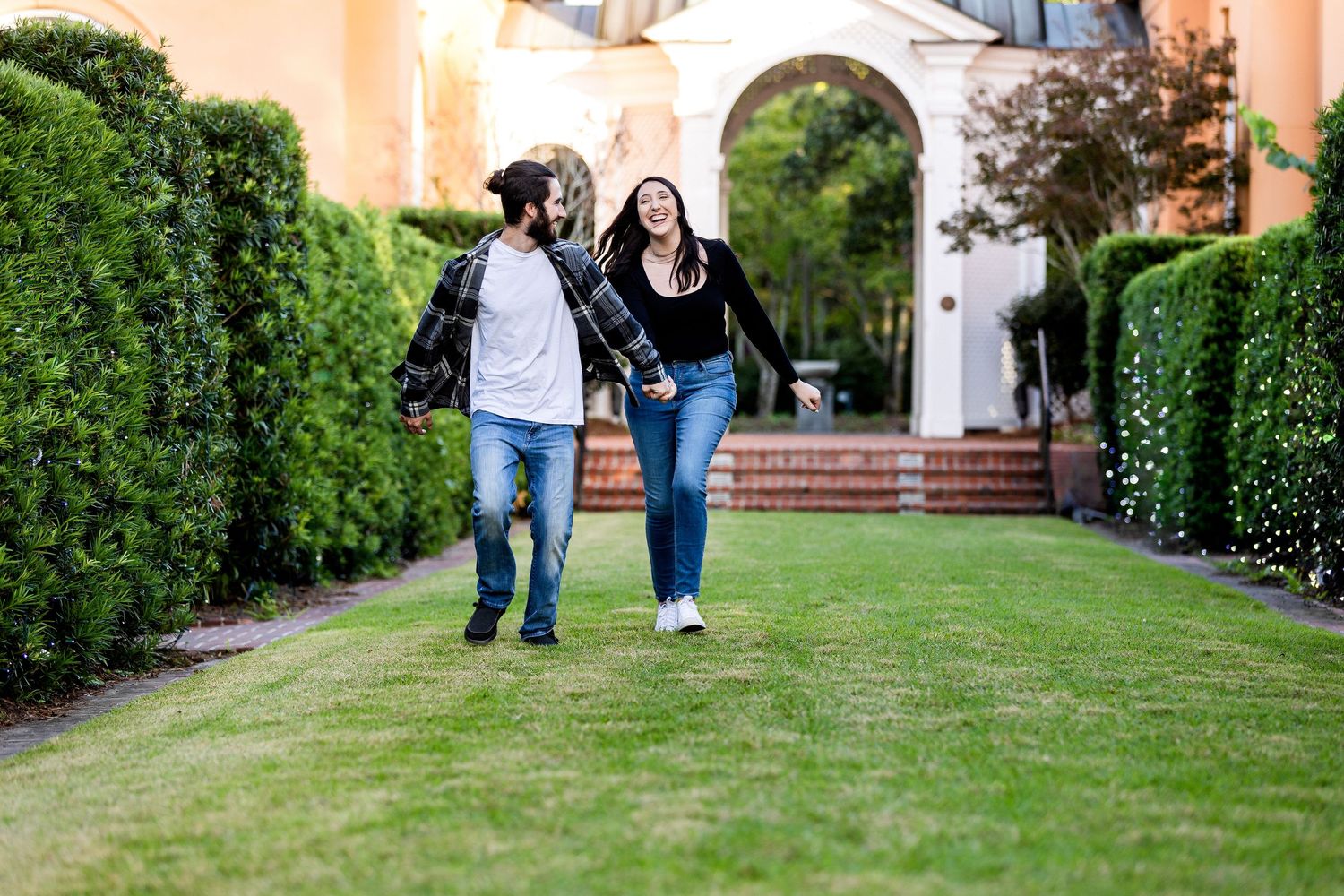 Two students walk together along a grass path bordered by hedges with arched architecture in the background.