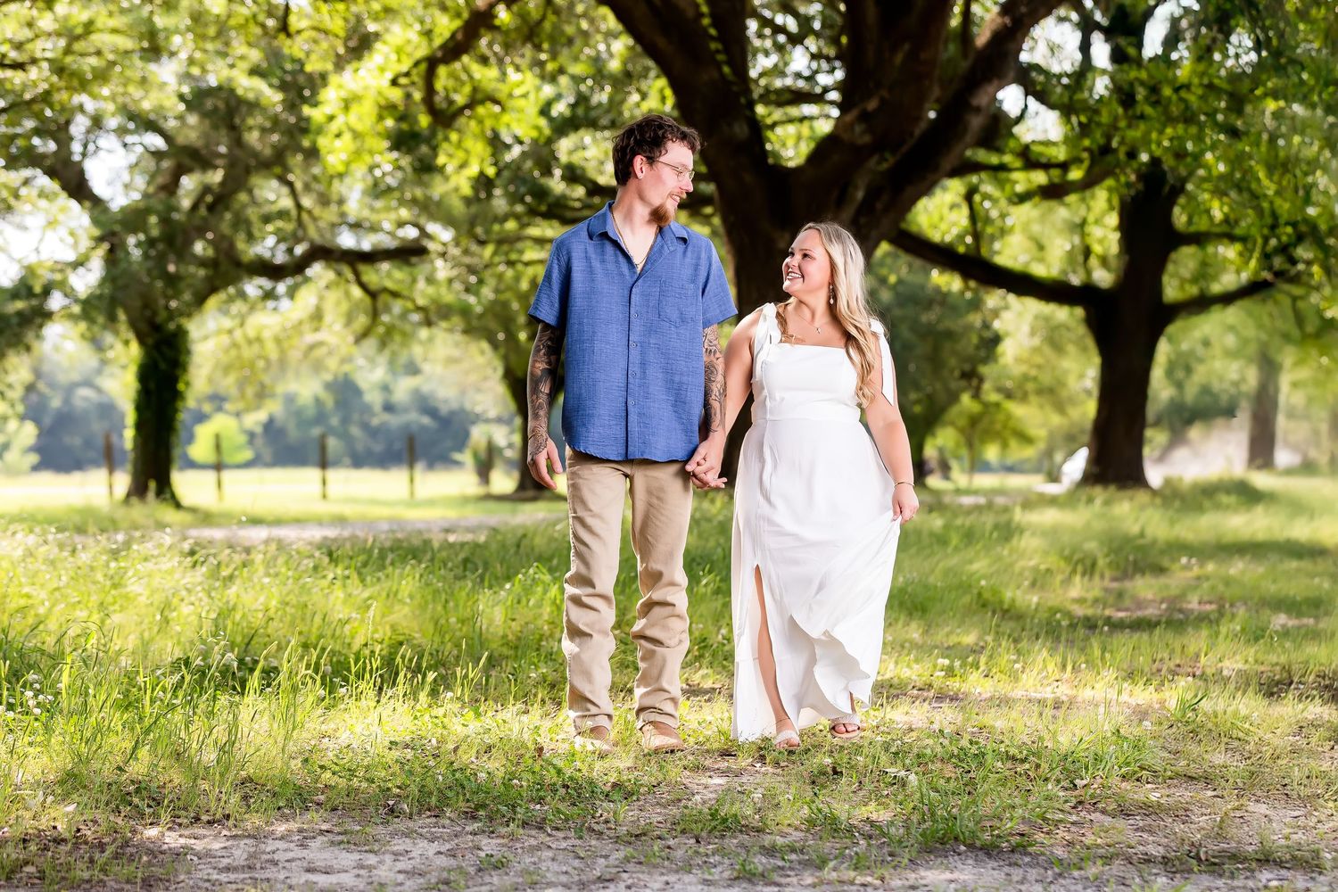 Couple walks hand in hand through a sunlit park with lush green trees and natural scenery.