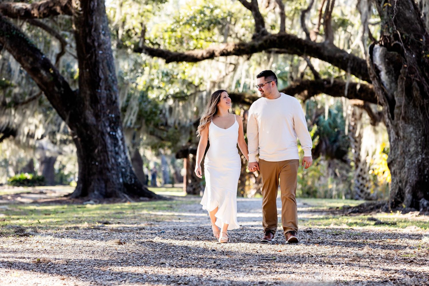 A couple walks hand in hand along a tree-lined path with Spanish moss hanging from above on a sunny afternoon.