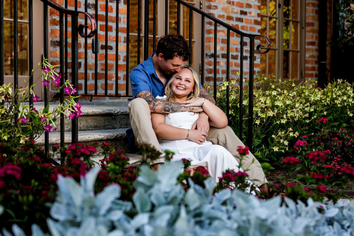 A couple shares an intimate moment sitting on steps surrounded by colorful flowers and foliage in a garden setting.
