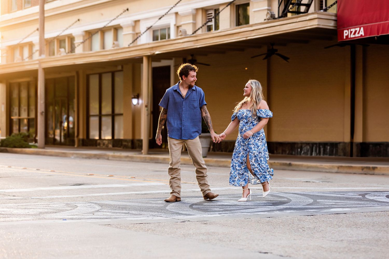A couple playfully dances together on a street with historic storefronts and warm evening light.