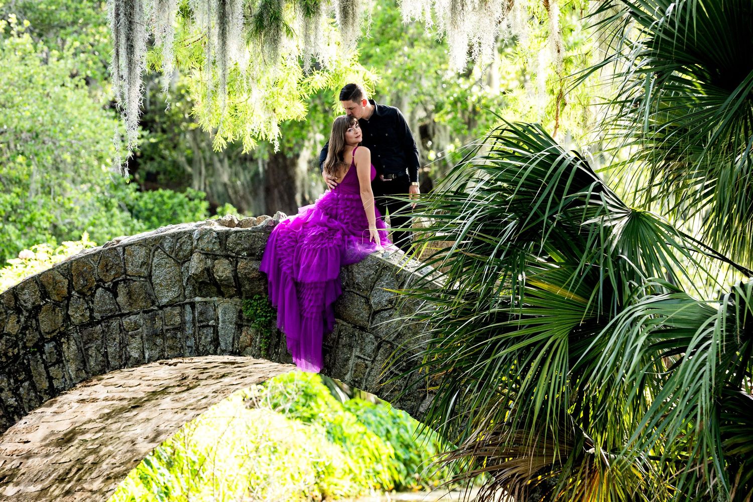 A person in a flowing purple dress walks along a stone bridge in a lush tropical garden setting.