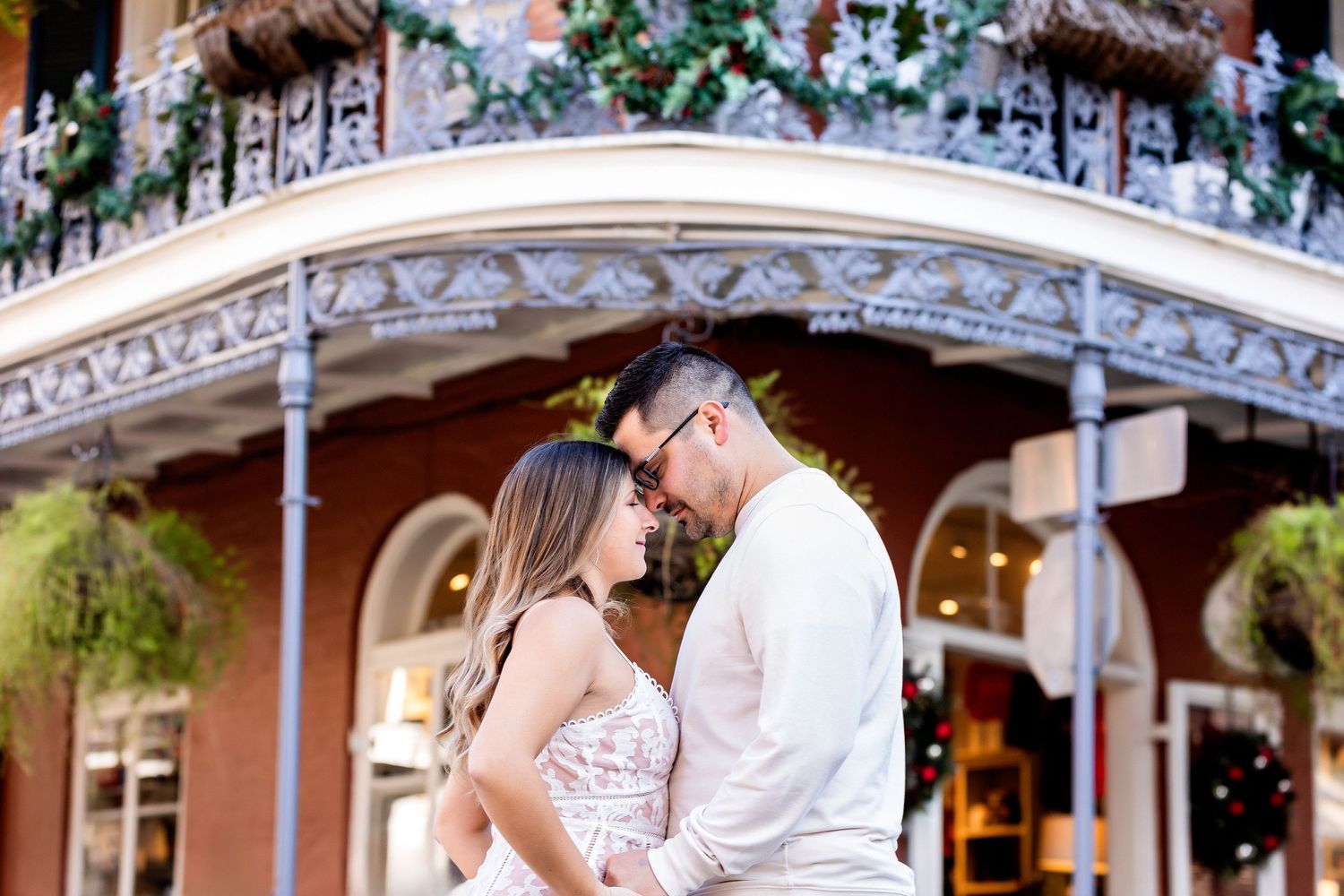 A romantic couple shares a tender moment under an ornate wrought iron balcony decorated with flowers in New Orleans.
