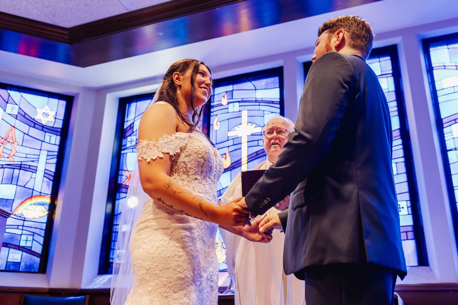 A couple exchanges vows during a wedding ceremony in a church with stained glass windows in the background.