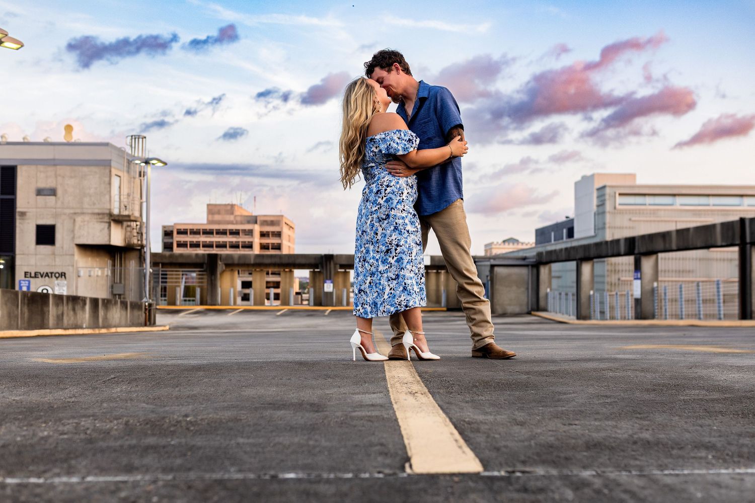 A romantic couple embraces on a wooden board on a rooftop parking deck at sunset with urban buildings in background.
