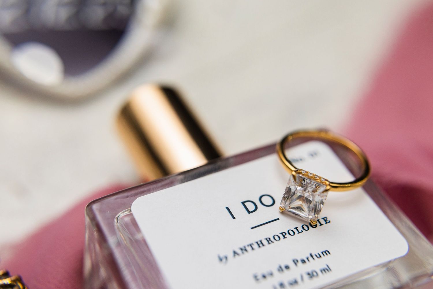 An engagement ring rests on a perfume bottle labeled 'I DO' against a soft pink background.