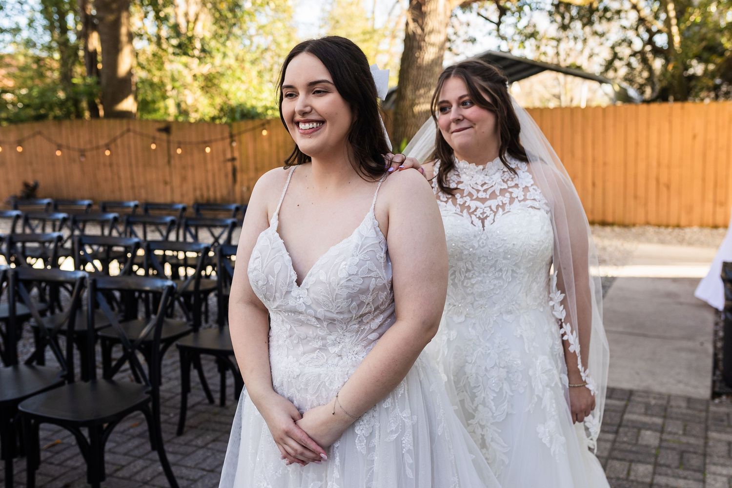 Three people in white lace wedding dresses pose together at an outdoor garden venue with string lights.