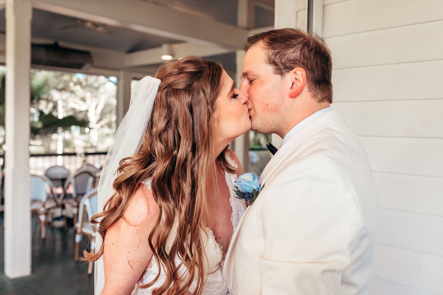 A couple shares their first kiss as newlyweds at a bright outdoor venue with the bride wearing a flowing veil.