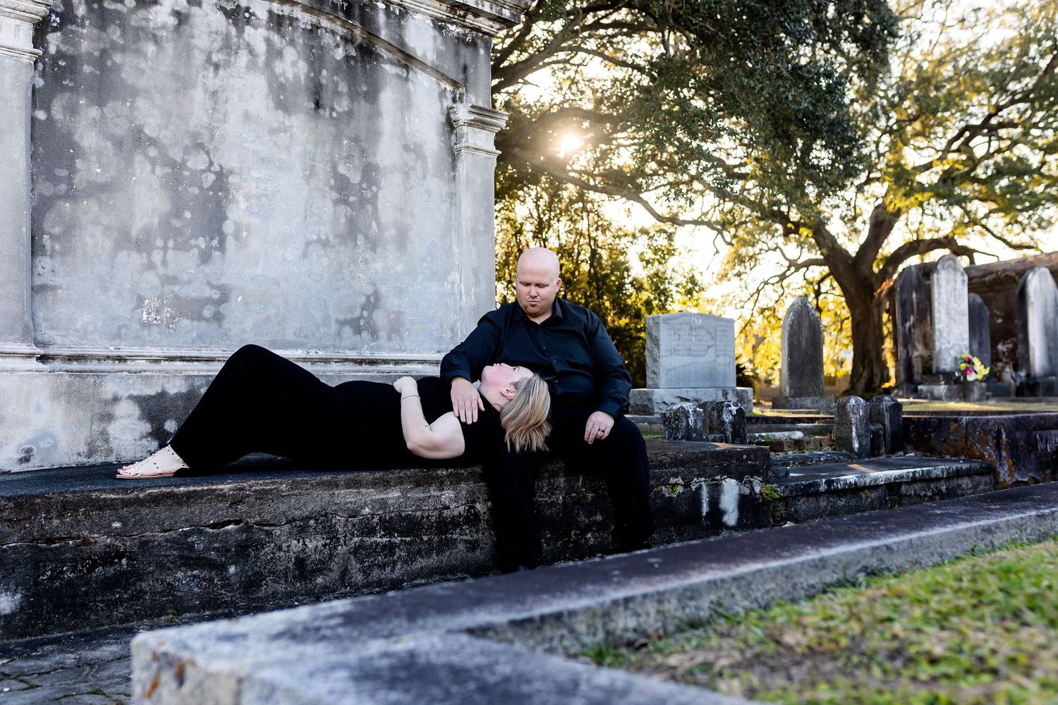 Two people in dark clothing pose on a stone ledge in a historic cemetery with sunlit trees in the background.