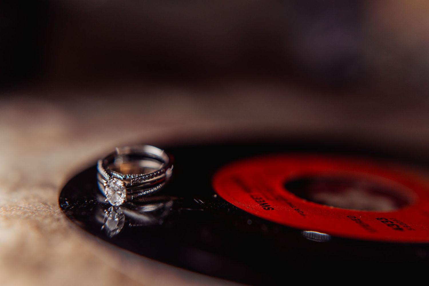 A sparkling diamond engagement ring rests on a red and black textured surface in dramatic lighting.