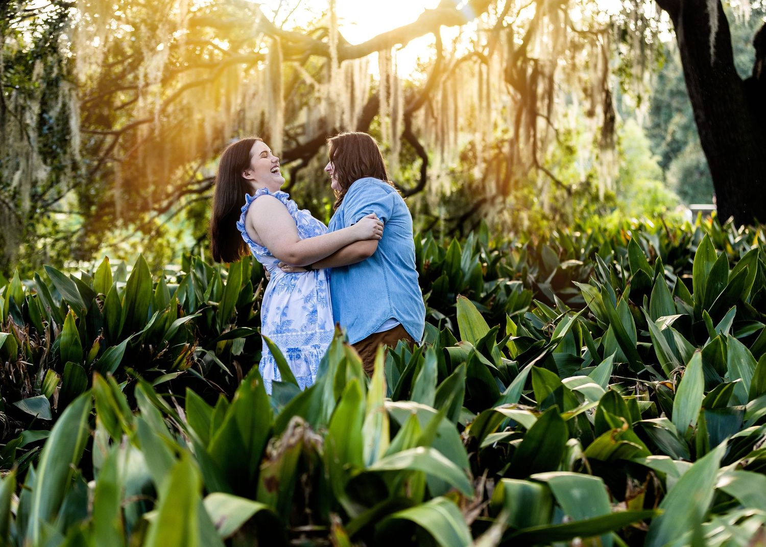 Two people share an intimate embrace in a lush tropical garden at sunset with light filtering through palm trees.