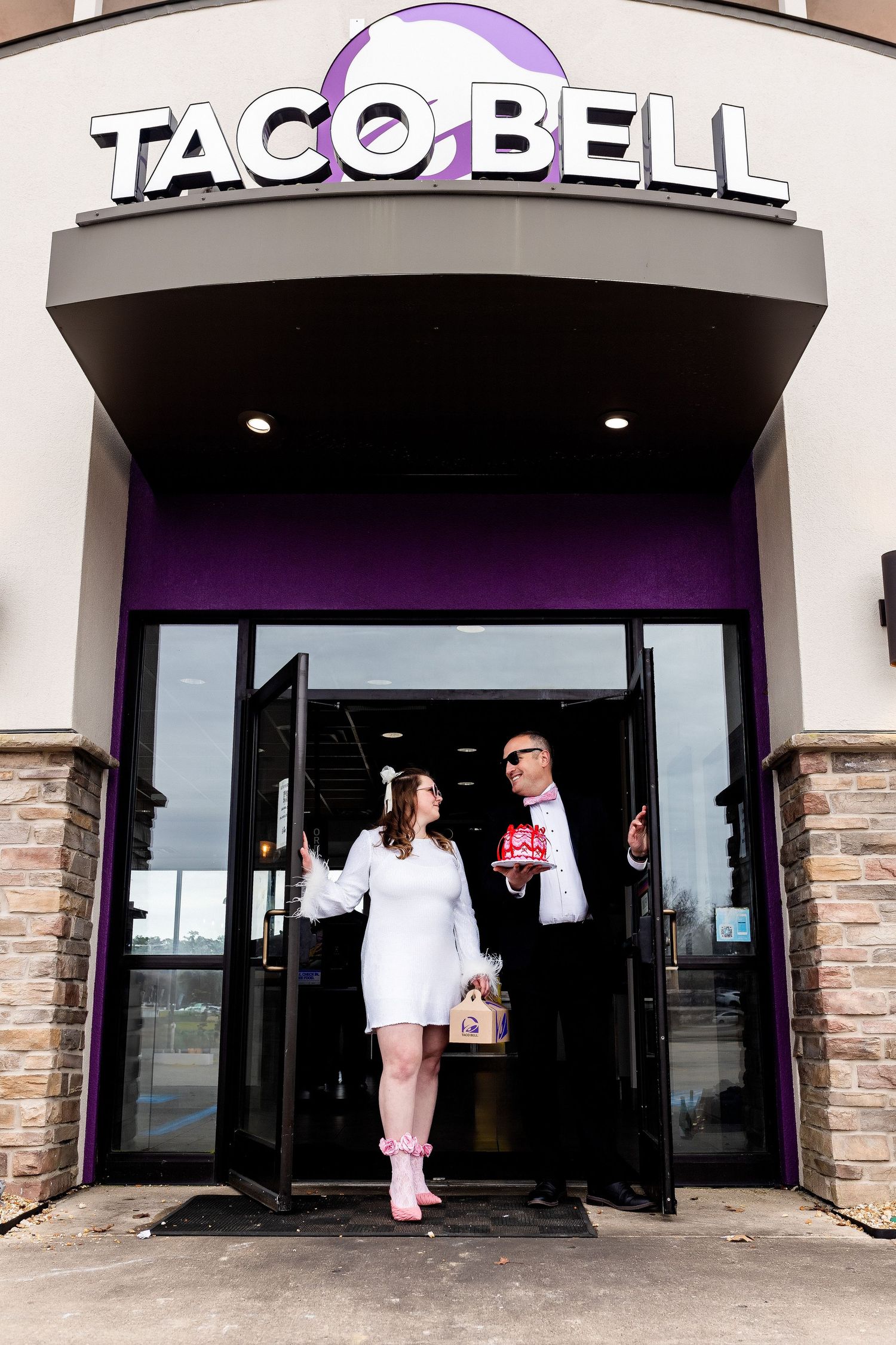 A couple poses in formal attire while exiting a Taco Bell restaurant under its purple and white illuminated storefront sign.