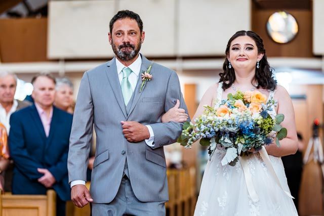 A wedding party processes down the aisle in elegant formal attire at an indoor ceremony.