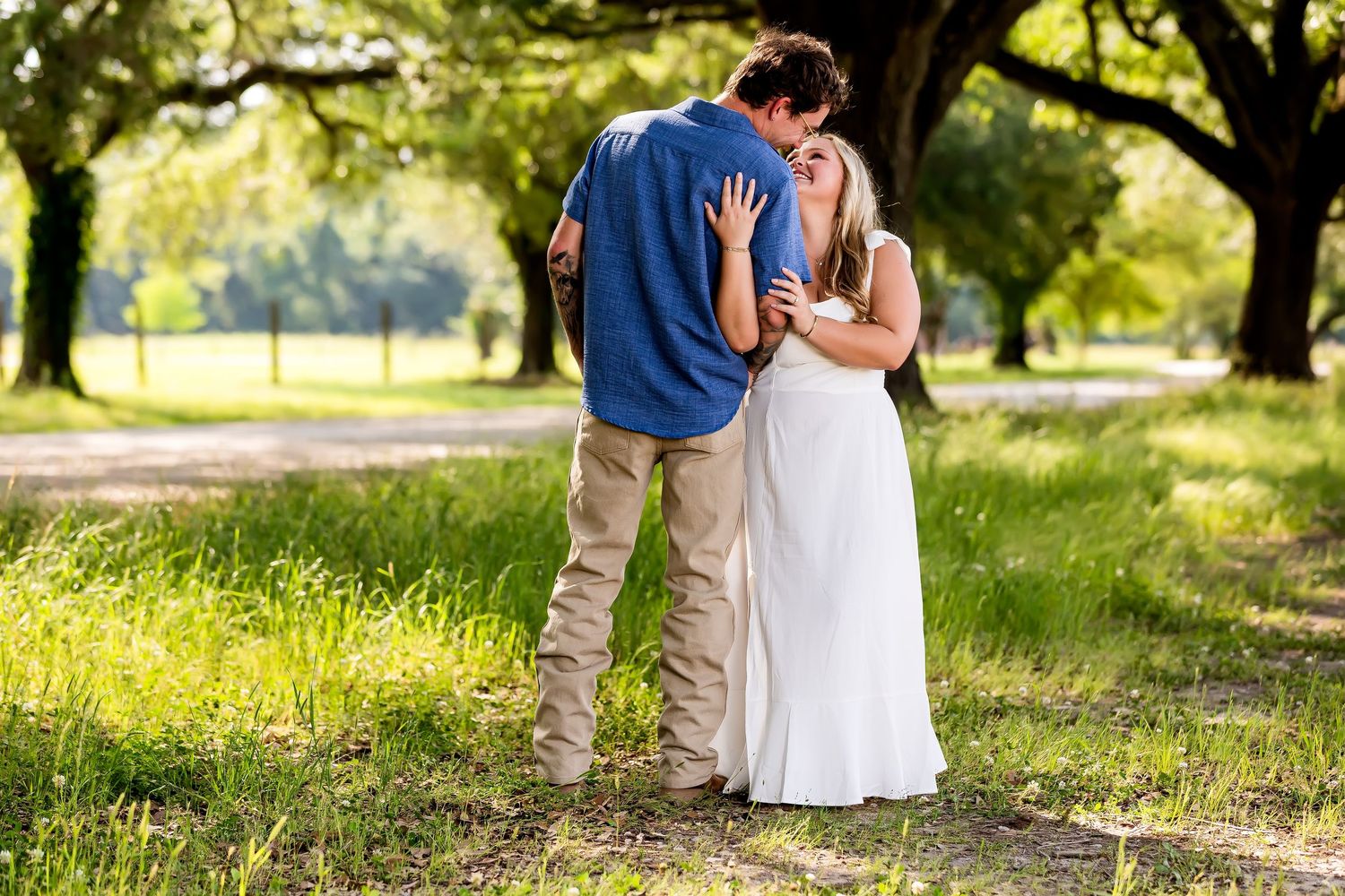 A couple embraces in a sunlit park setting with green trees and golden light filtering through.