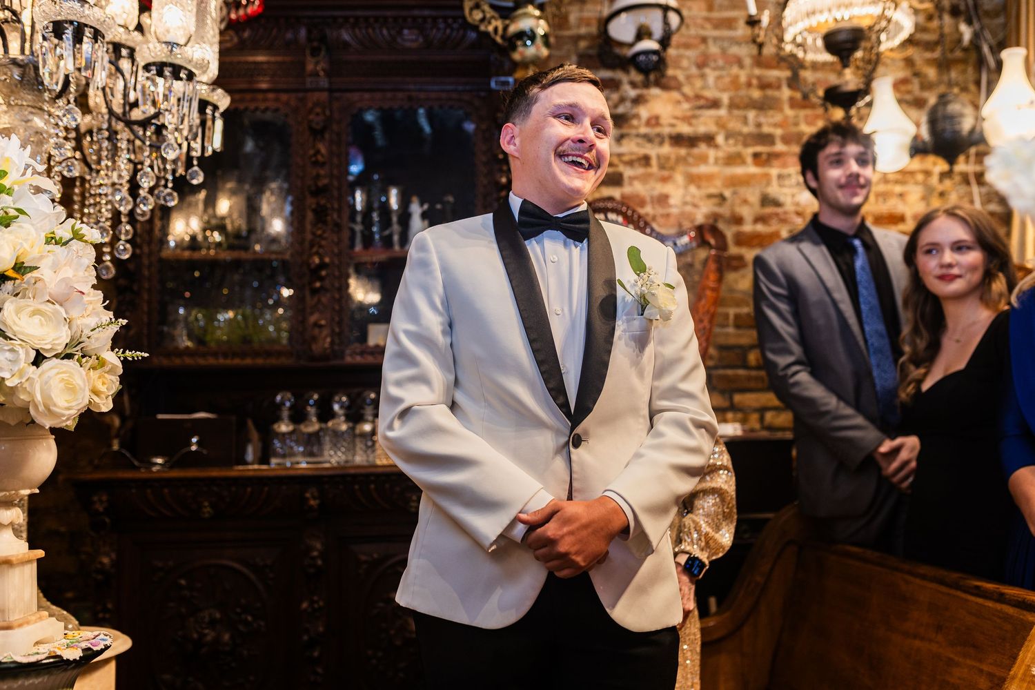 A person in a white dinner jacket and bowtie laughs during an elegant event with brick walls and chandeliers.