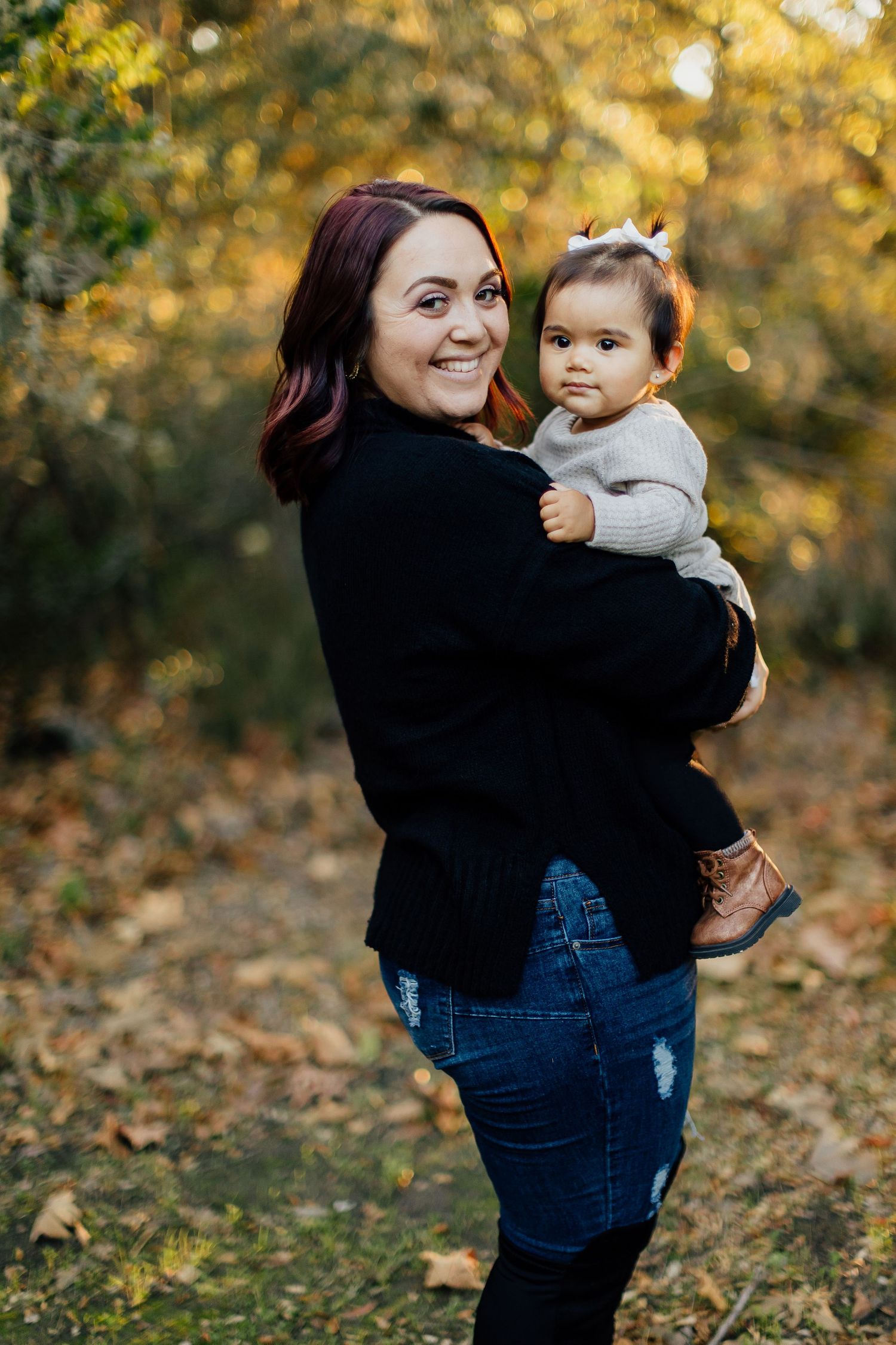 An adult and small child pose together outdoors with autumn foliage in the background during a fall photo session.