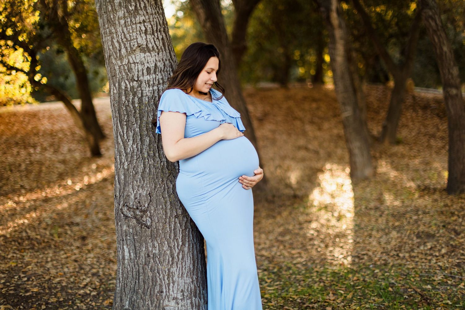 Expecting mother in flowy light blue dress stands thoughtfully in a sunlit forest setting.