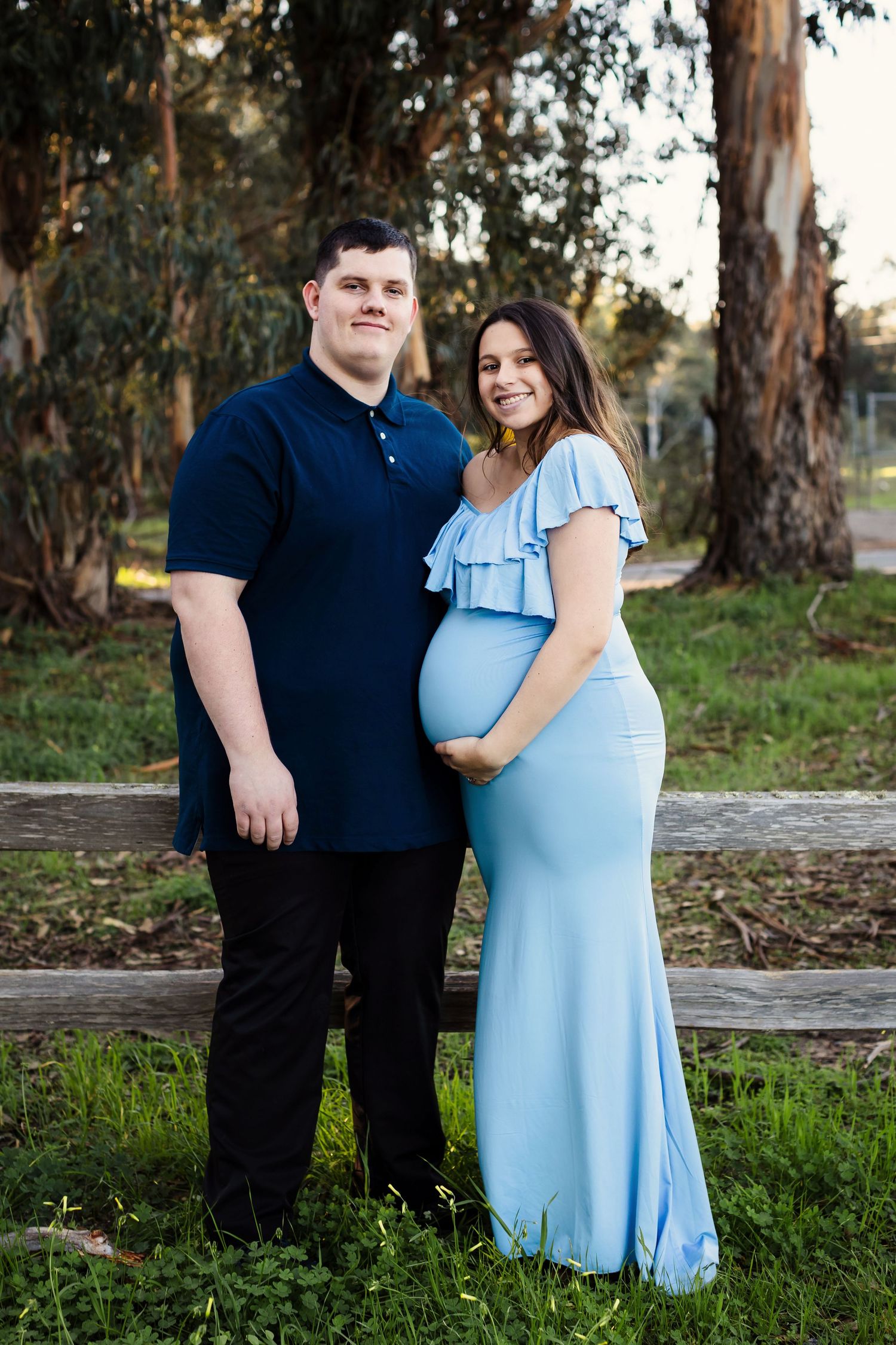 A couple in coordinated light blue and navy outfits poses outdoors on green grass near trees at sunset.