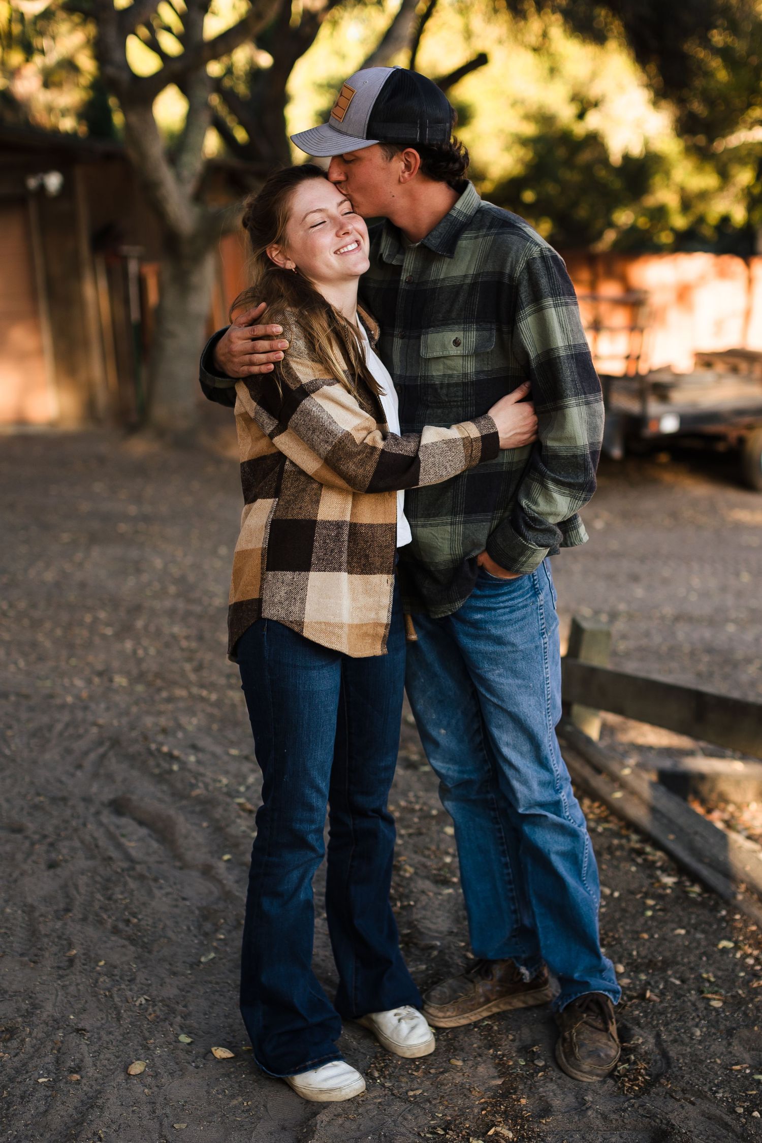 A couple stands close together under golden sunlight wearing cozy fall attire including a plaid scarf and flannel shirt.
