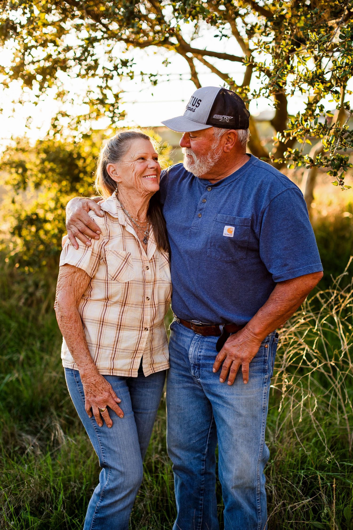 A loving mature couple embraces while standing outdoors in warm sunset lighting beneath tree branches.