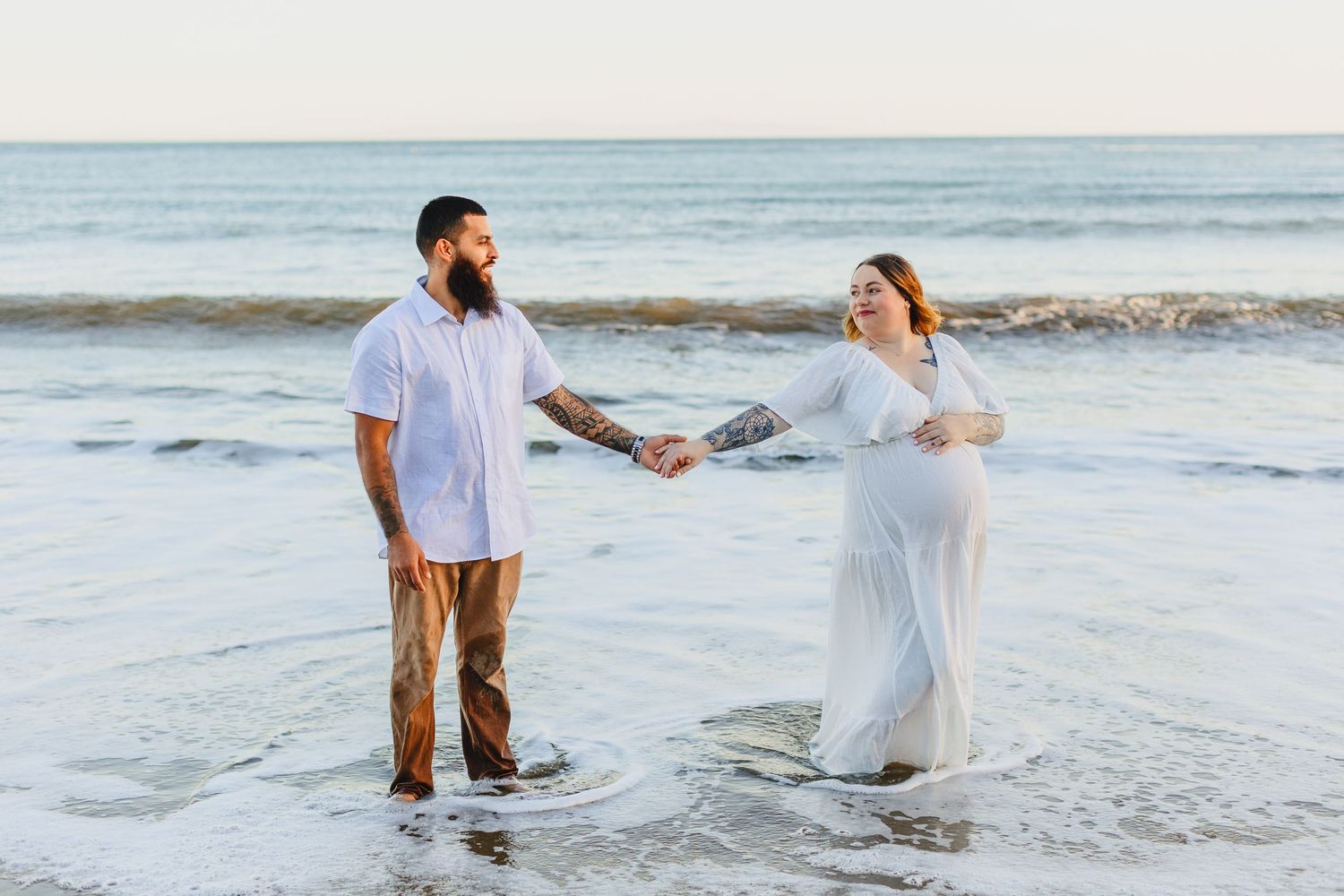 A couple stands together holding hands on a beach at sunset, with waves gently rolling in behind them.