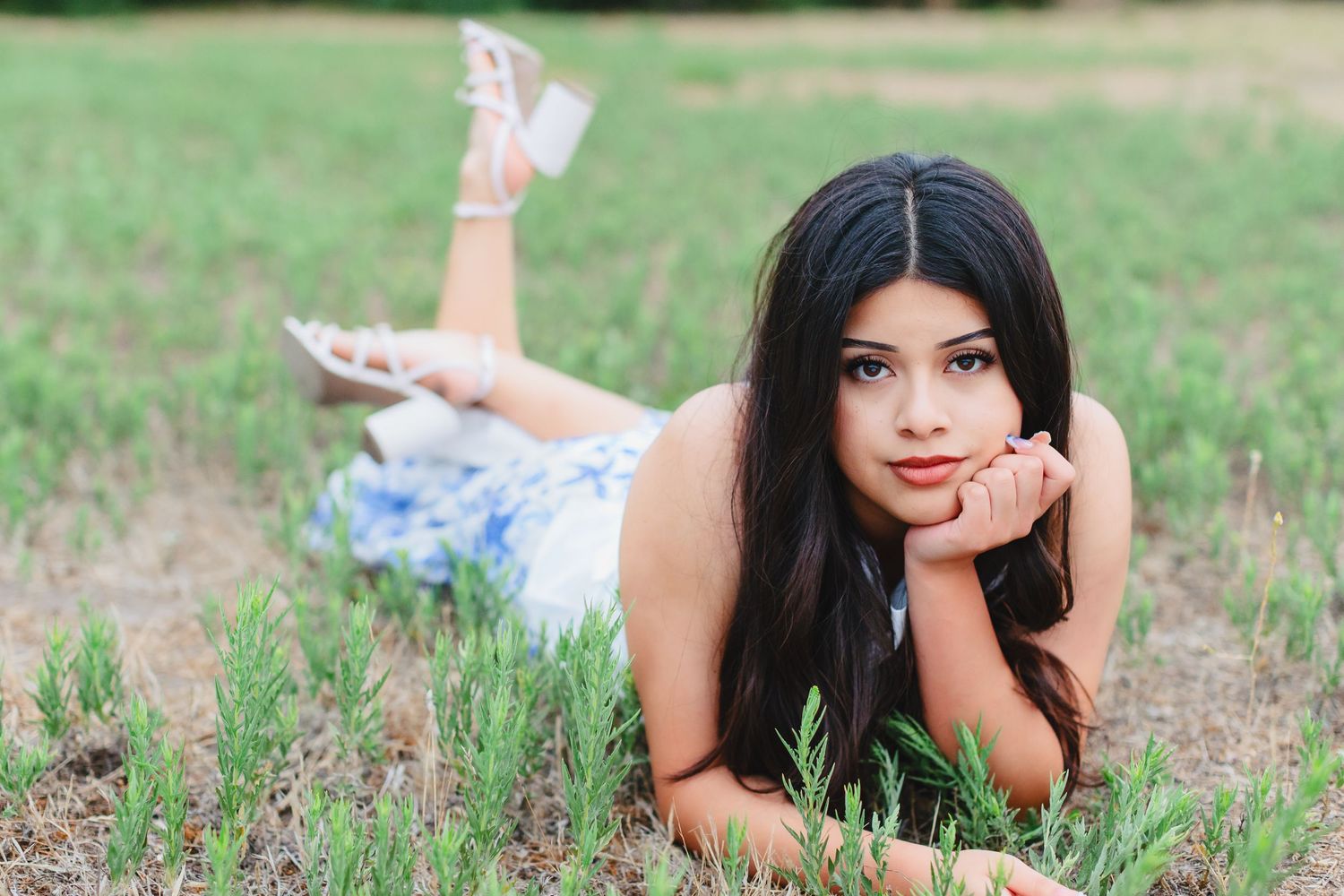 A person with long dark hair lies in a grassy field wearing a blue dress while resting on their elbows.