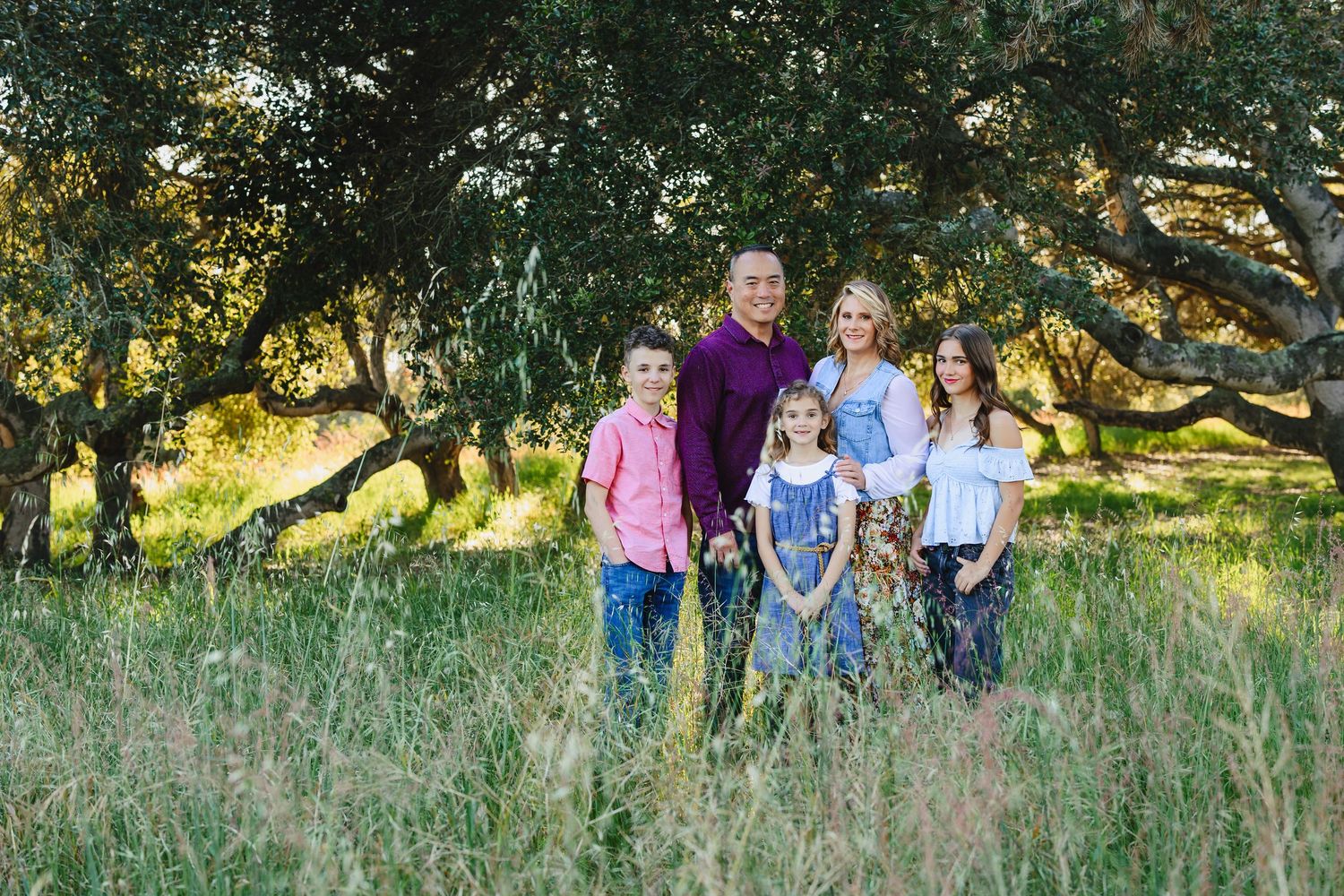 Family of six posing outdoors in a lush green meadow surrounded by trees during golden hour.
