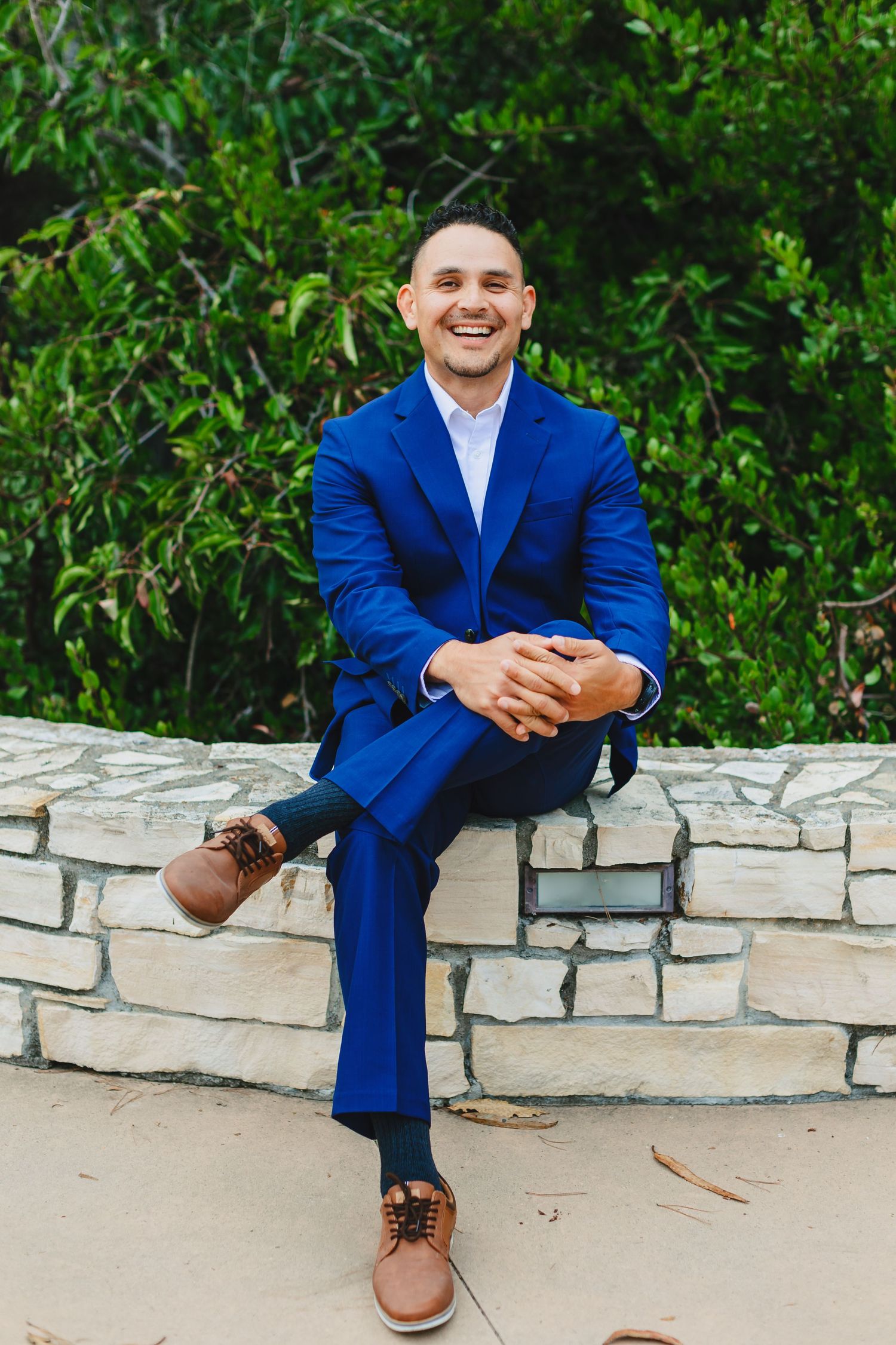 Professional seated on stone wall wearing bright blue suit with brown shoes in outdoor setting.